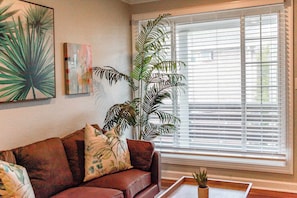 Una sala de estar con un sofá de cuero marrón, una planta grande en maceta y una ventana con persianas.