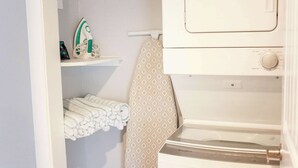 A laundry area with a washing machine, an iron, and neatly folded towels on a shelf. - Sojourn Palm Suite at Beach Cottages (Norfolk)