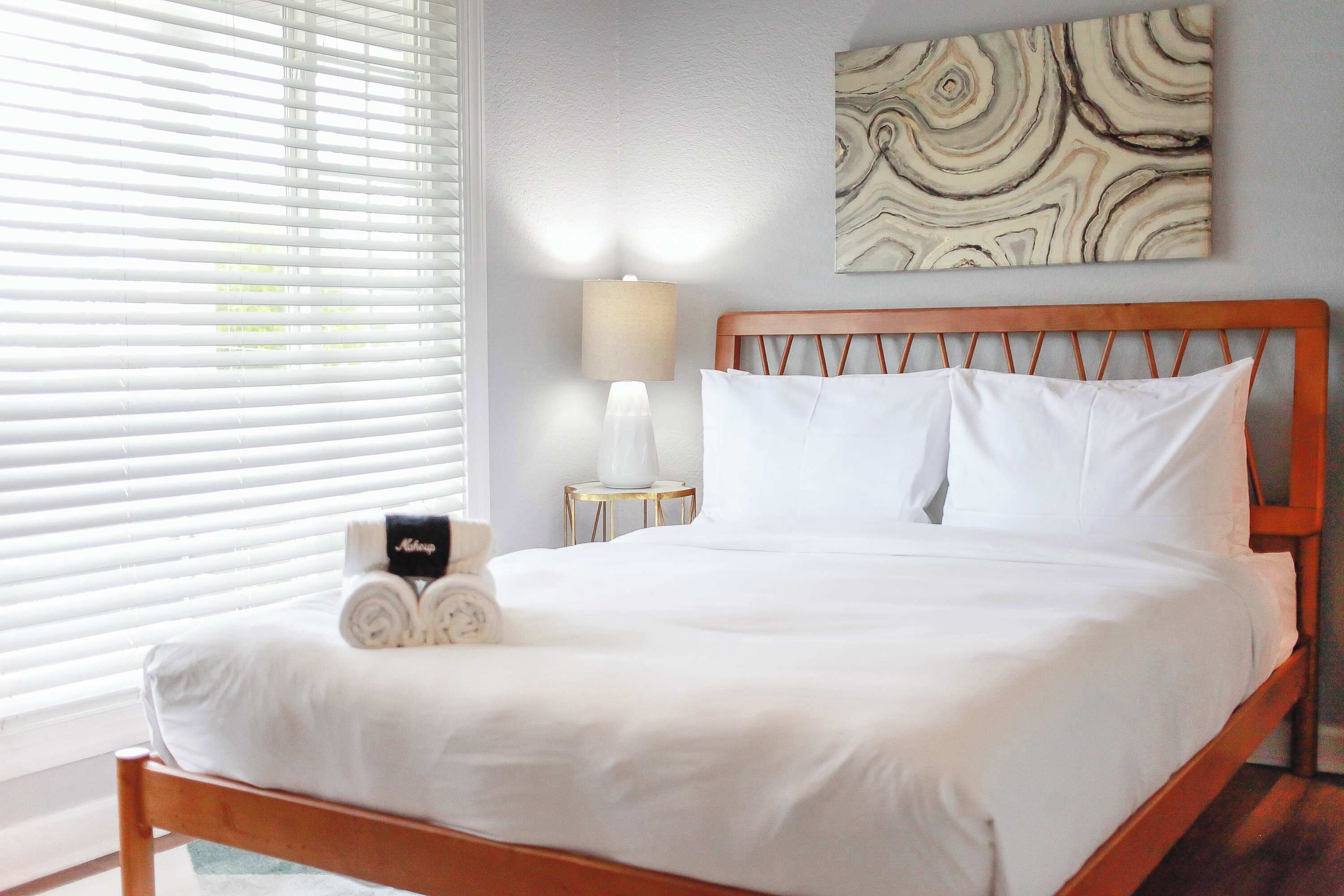 Uma cama bem arrumada com lençóis brancos, cabeceira de madeira e uma obra de arte abstrata pendurada na parede.