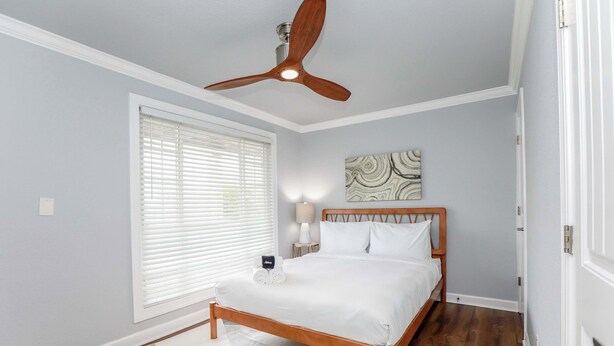 A bedroom with a wooden bed, a ceiling fan, a window with blinds, a bedside lamp, and a wall art piece. - Sojourn Palm Suite at Beach Cottages (Norfolk)