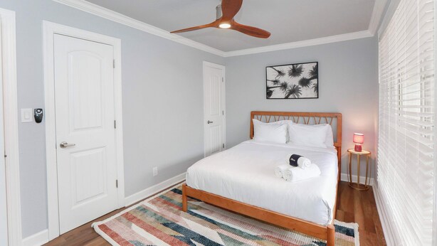 A bedroom with a wooden bed, white walls, a ceiling fan, a door, a framed picture, a bedside lamp, and a striped rug. - Sojourn Beach Suite 2 BR 2 BA at Beach Cottages (Norfolk)