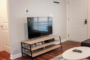 Ein Wohnzimmer mit einem Flachbildfernseher auf einem Holzständer, einem weißen Couchtisch und einer Tür im Hintergrund.