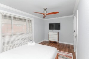 Un dormitorio con cama, ventilador de techo, televisor, cómoda y ventana con persianas.