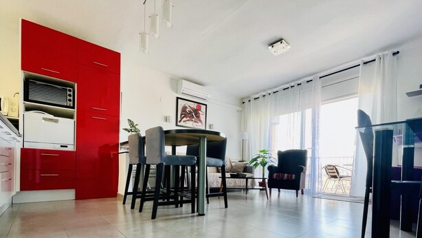 Un espace de vie moderne avec une cuisine rouge, un coin repas comprenant une table et des chaises, et un balcon avec un fauteuil.