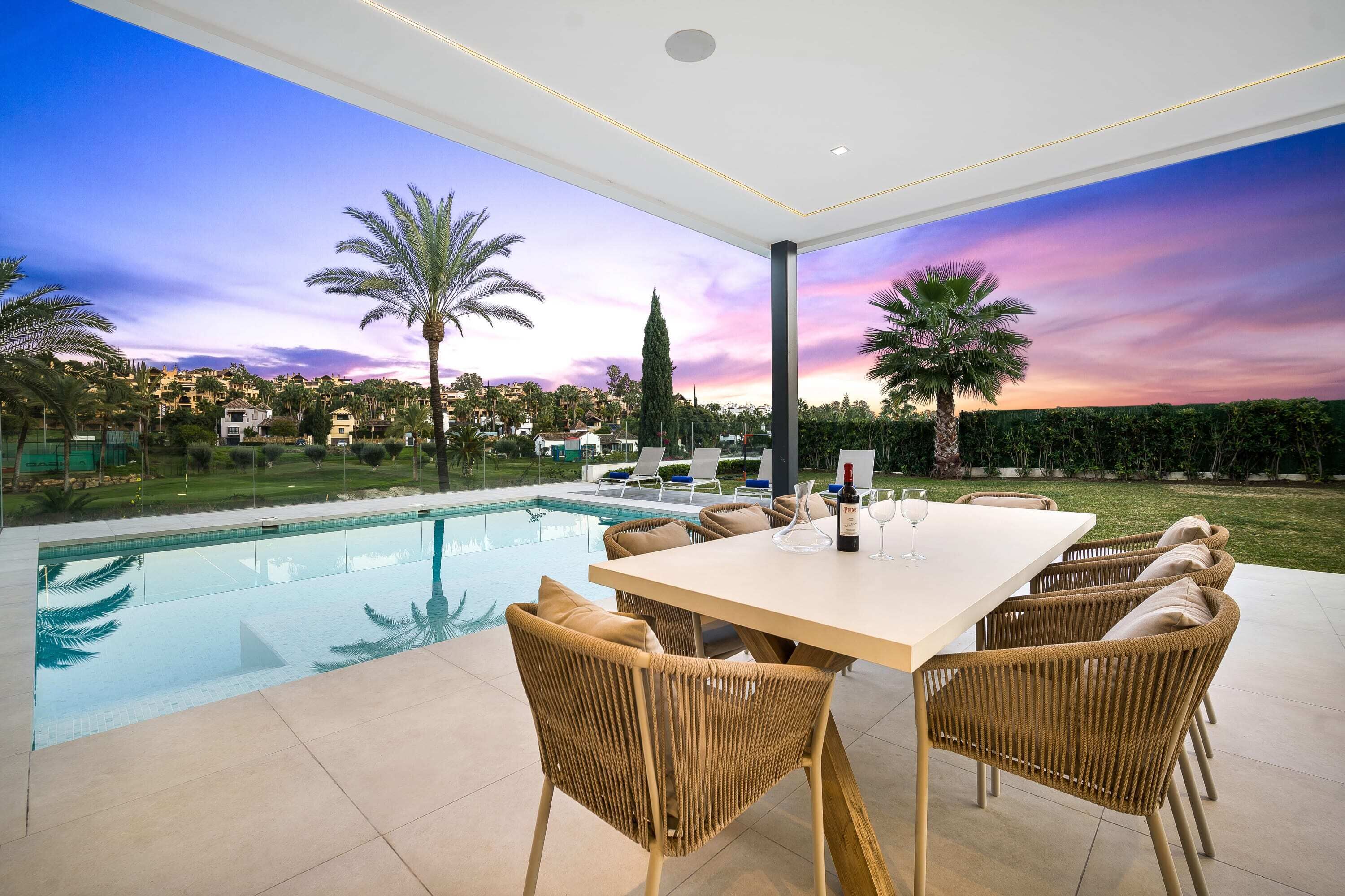 A poolside dining area with a table and chairs, a view of the pool, and a sunset in the background.