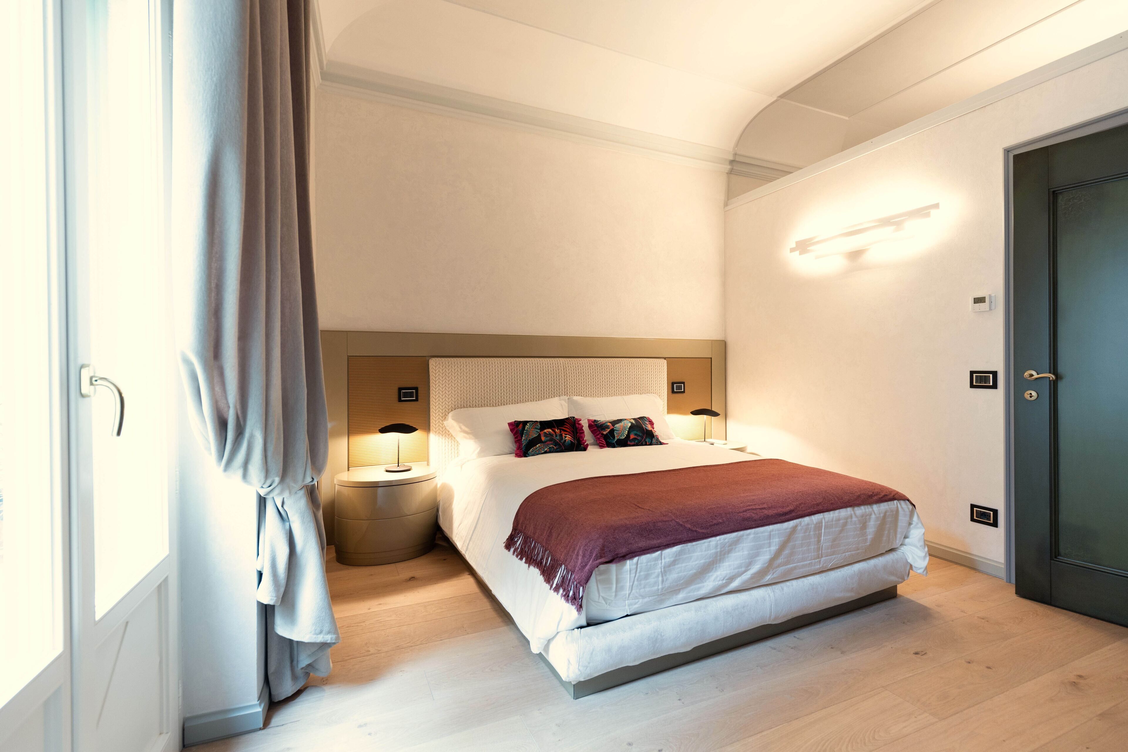 Un dormitorio con cama, lámparas de noche, mesita de noche, una puerta y cortina.