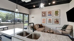 Una cocina moderna con encimeras de granito, un lavamanos doble y lavavajillas. Hay una zona de comedor con un banco y un sofá. La habitación tiene ventanas amplias con vistas a coches aparcados y árboles.
