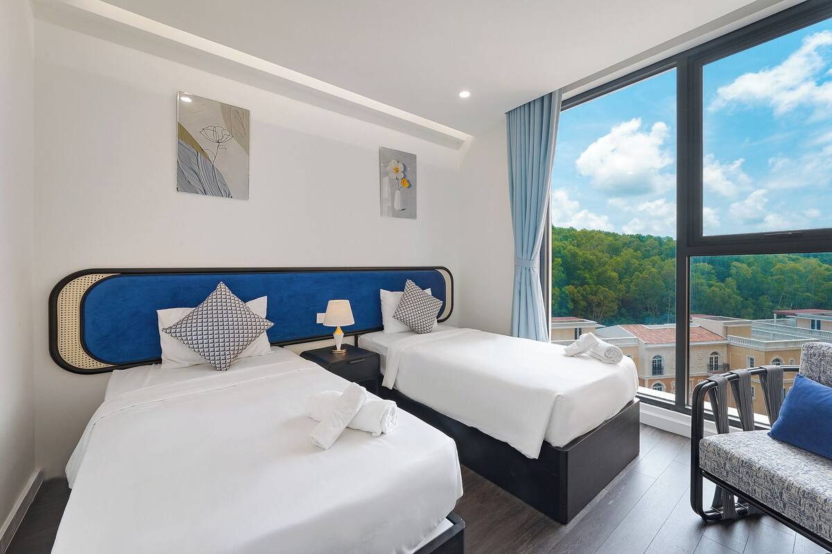 A hotel room with two beds, a large window with a city view, and artwork on the walls.