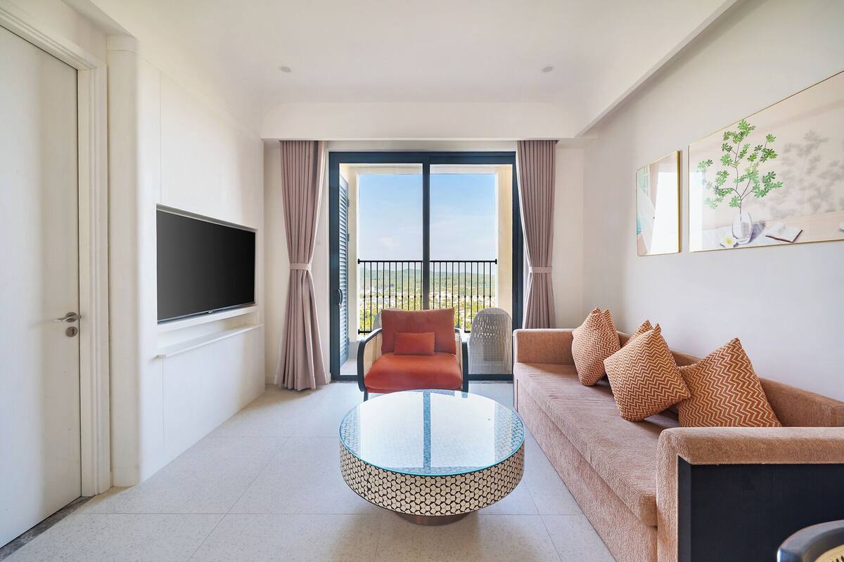 A modern living room with a round glass coffee table, a sofa with patterned cushions, a flat-screen TV mounted on the wall, and a balcony with a view.