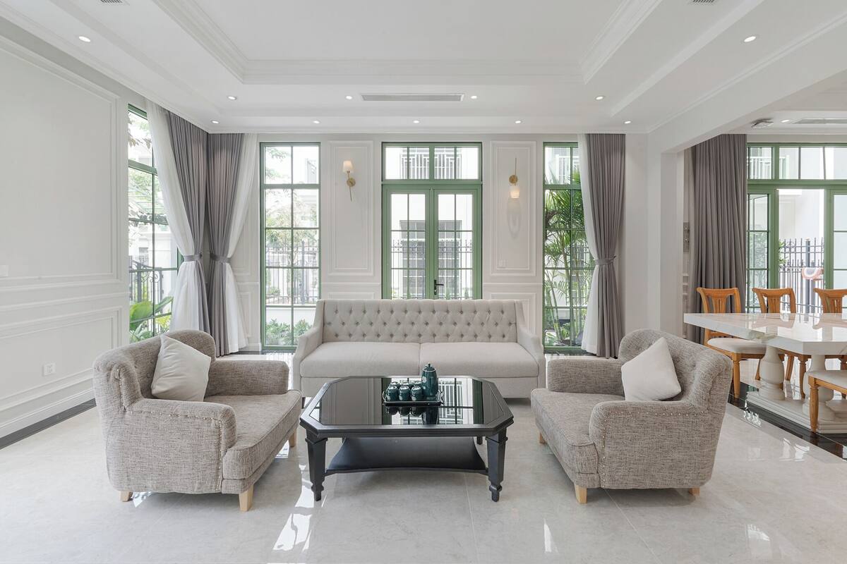 A spacious living room with a beige sofa, two armchairs, a glass coffee table, and a dining area with a table and chairs.