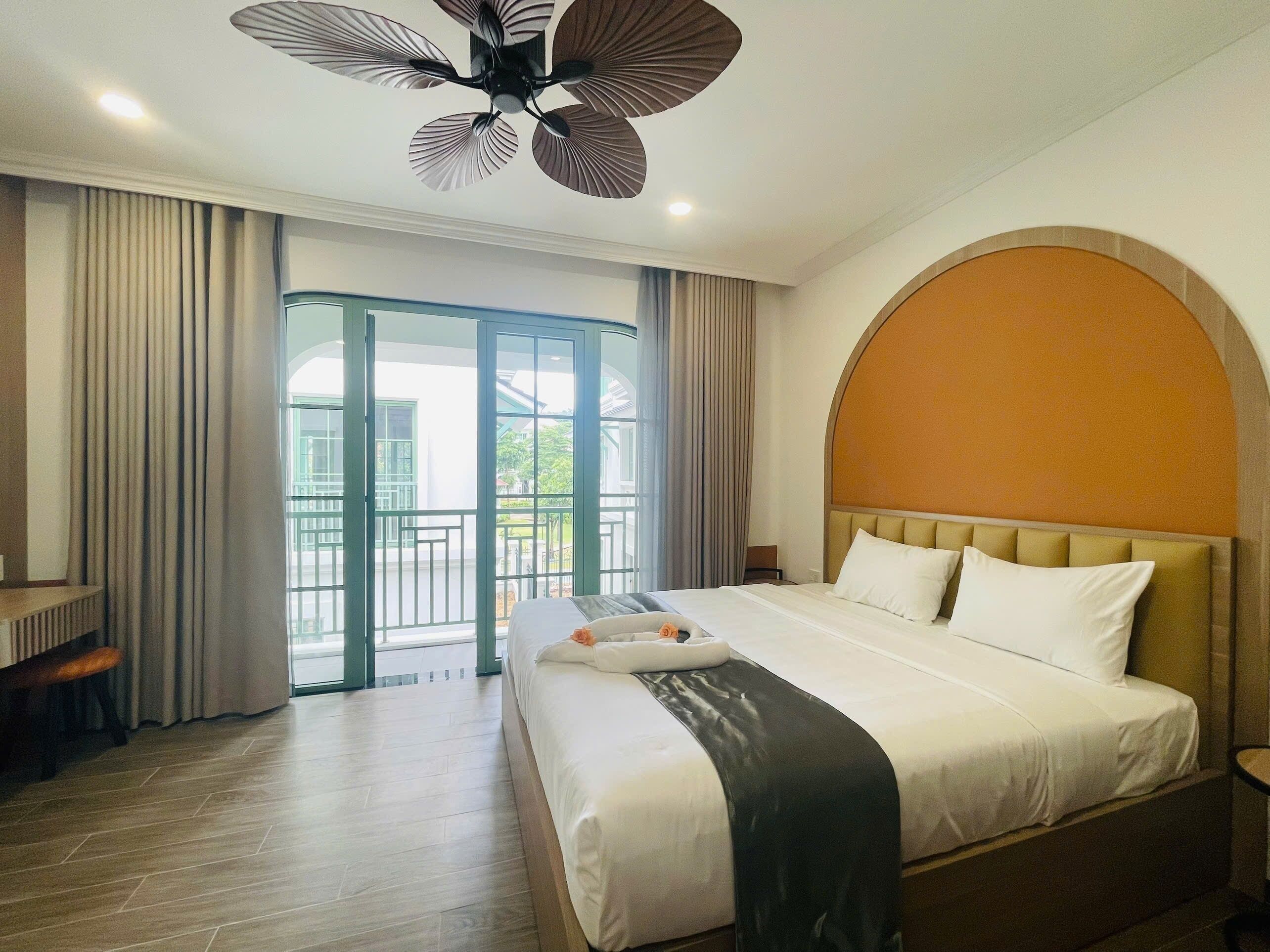A hotel room with a bed, a desk, a chair, a ceiling fan, and a view of the outdoors.