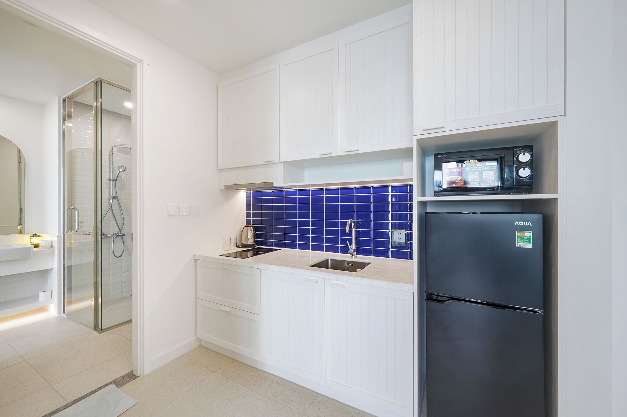 A modern kitchen with white cabinetry, a blue-tiled backsplash, a stainless steel refrigerator, and a glass-enclosed shower area.