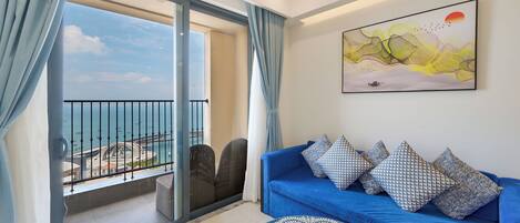 A balcony with a view of the sea, a blue sofa with patterned cushions, and a wall-mounted abstract painting.