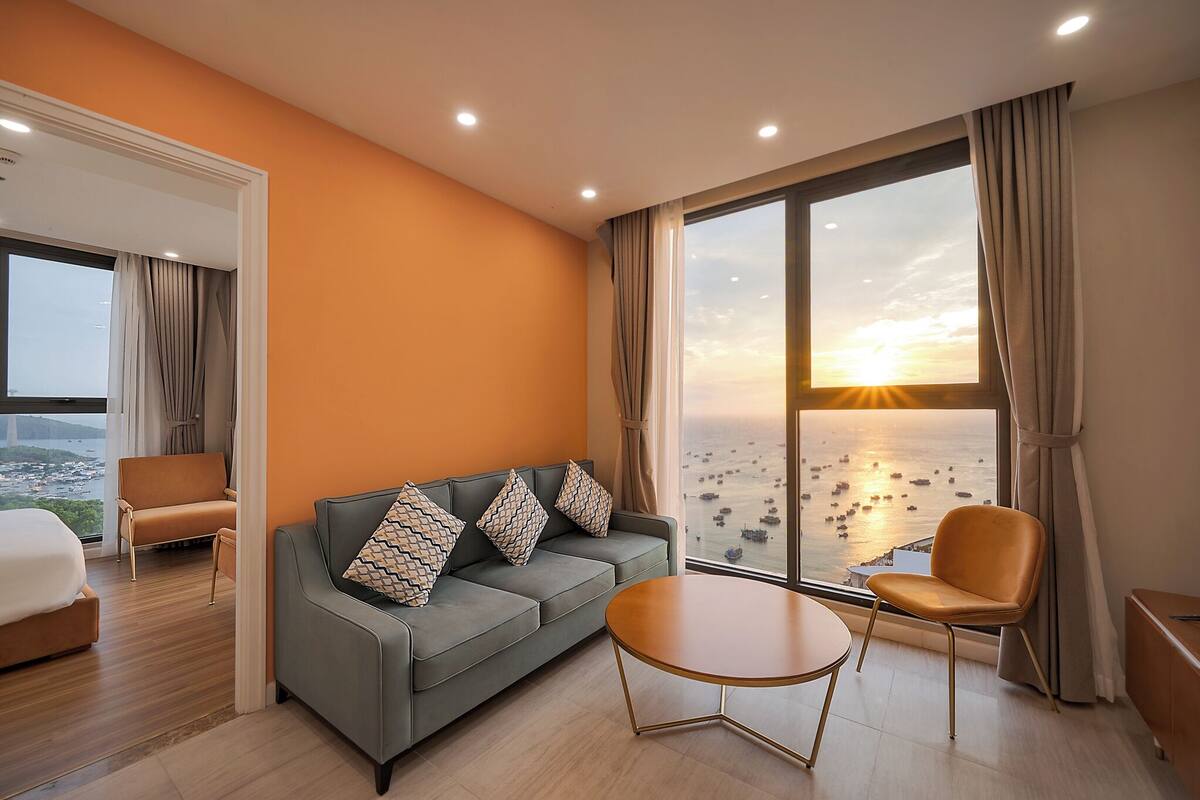 A modern hotel room with a large window offering a view of the sea and boats, a grey sofa with patterned cushions, a wooden coffee table, and a bed with a white headboard.