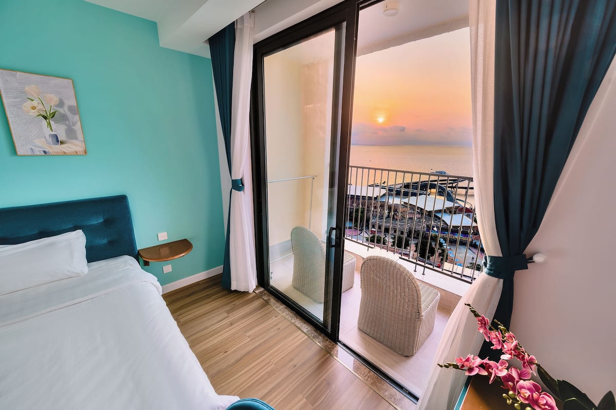 A hotel room with a bed, a chair, a balcony with a view of the sea, and a painting on the wall.