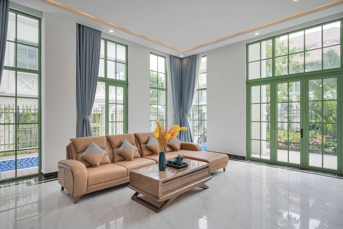 A spacious living room with a beige sectional sofa, a marble coffee table, and large windows with green frames.