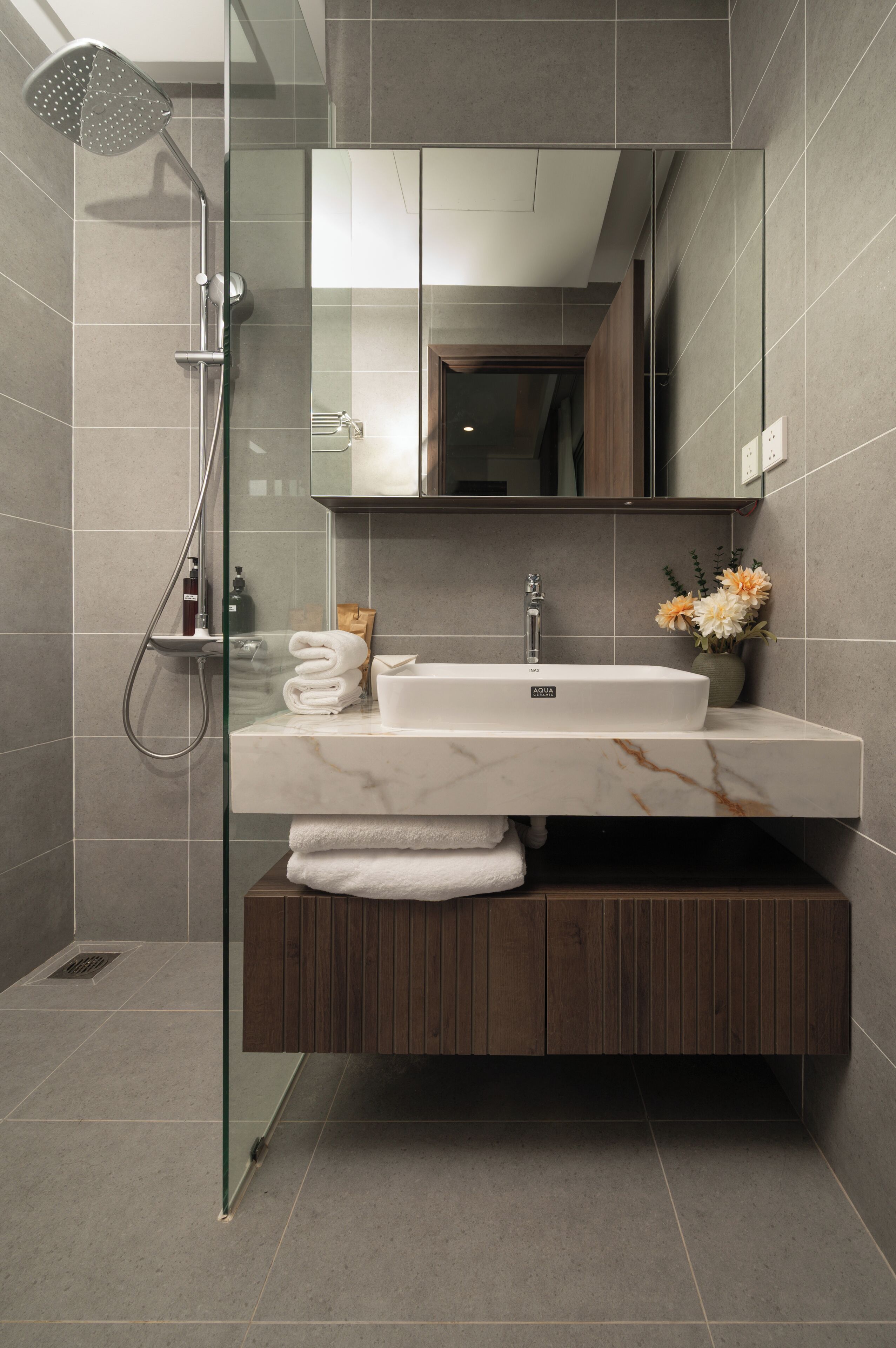 Un baño moderno con cabina de ducha de vidrio, un lavabo blanco con encimera de mármol y un mueble de tocador de madera.