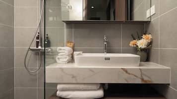 Un baño moderno con cabina de ducha de vidrio, un lavabo blanco con encimera de mármol y un mueble de tocador de madera.