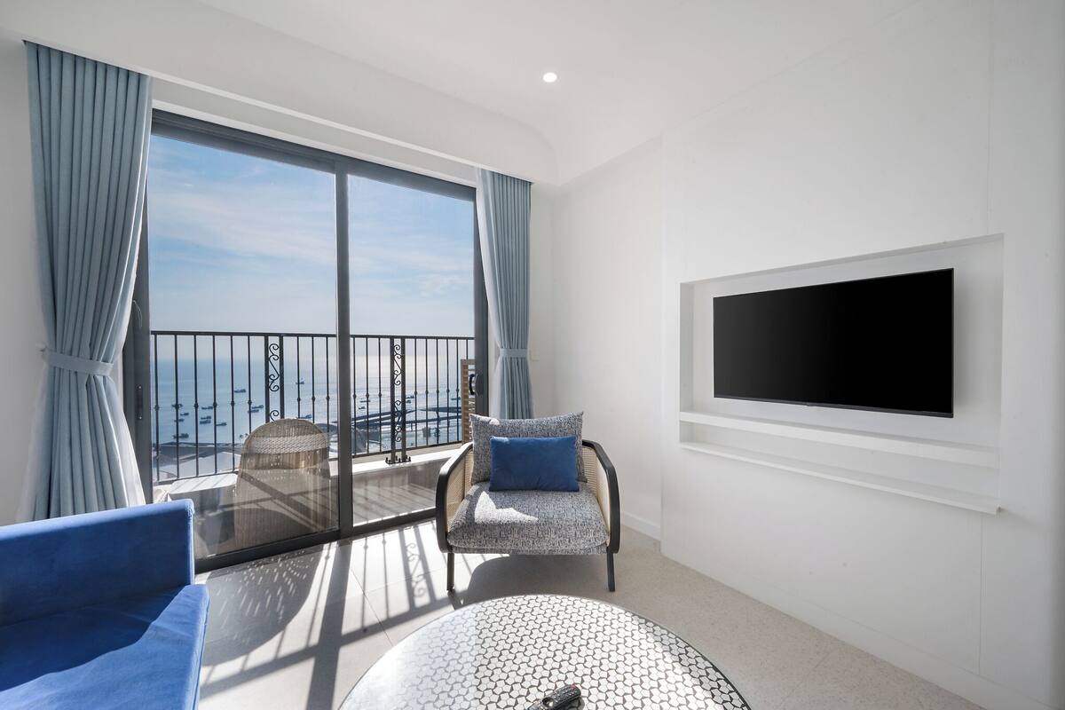 A balcony with a view of the sea, a TV mounted on the wall, and two chairs with cushions.