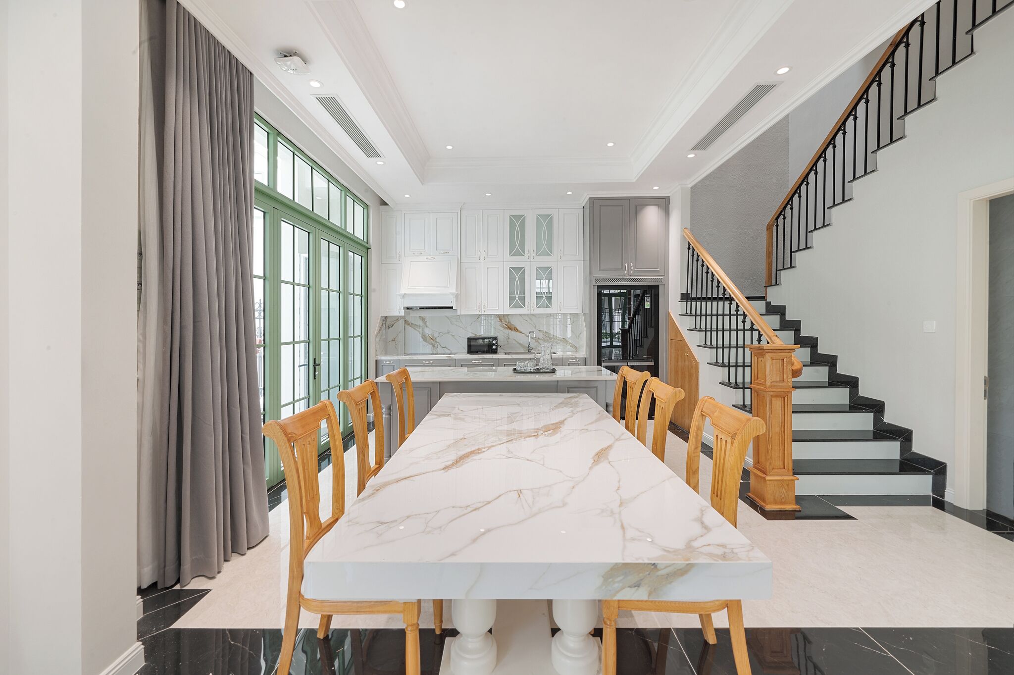 A spacious dining area with a marble table and wooden chairs, a kitchen with white cabinets, and a staircase with a wooden handrail.