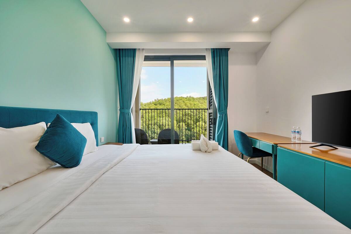 A hotel room with a bed, a desk, a chair, a TV, and a view of greenery through the window.