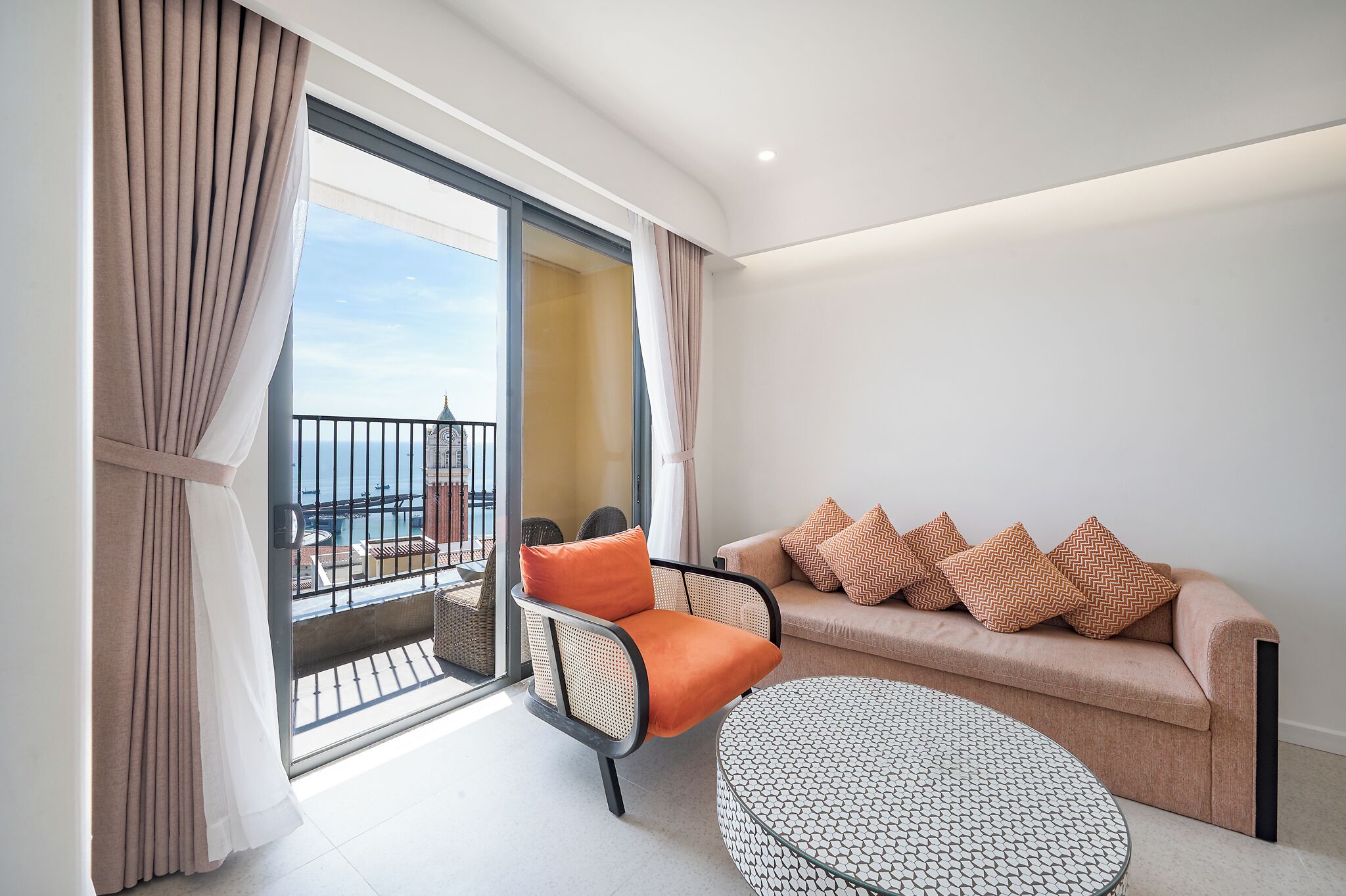 Un salon moderne avec un canapé, des coussins orange, une table basse ronde et un balcon doté d’une balustrade.