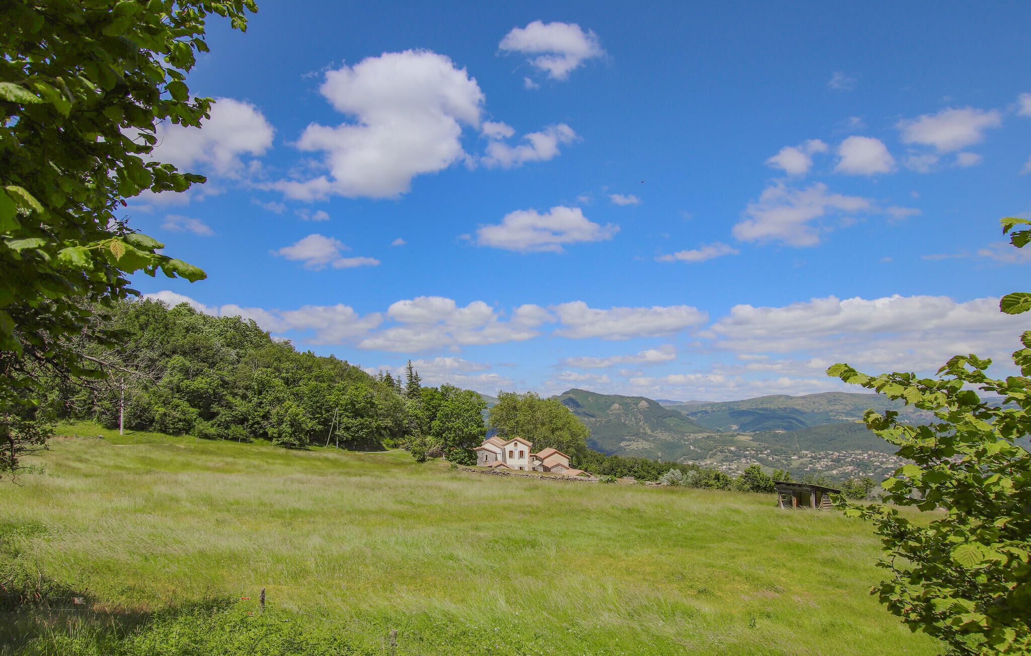 Ein grasbewachsenes Feld mit einem Haus und einer kleinen Struktur in der Ferne, umgeben von Bäumen und Hügeln.