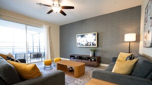 A modern living room with a sofa, coffee table, and a TV stand. There is a ceiling fan and a large window with a view of the outdoors. - 6BD Storey Lake Pool Home with Resort Amenities near DISNEY (Kissimmee)