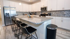A modern kitchen with white cabinets, a granite countertop, stainless steel appliances, and a central island with bar stools. - 6BD Storey Lake Pool Home with Resort Amenities near DISNEY (Kissimmee)