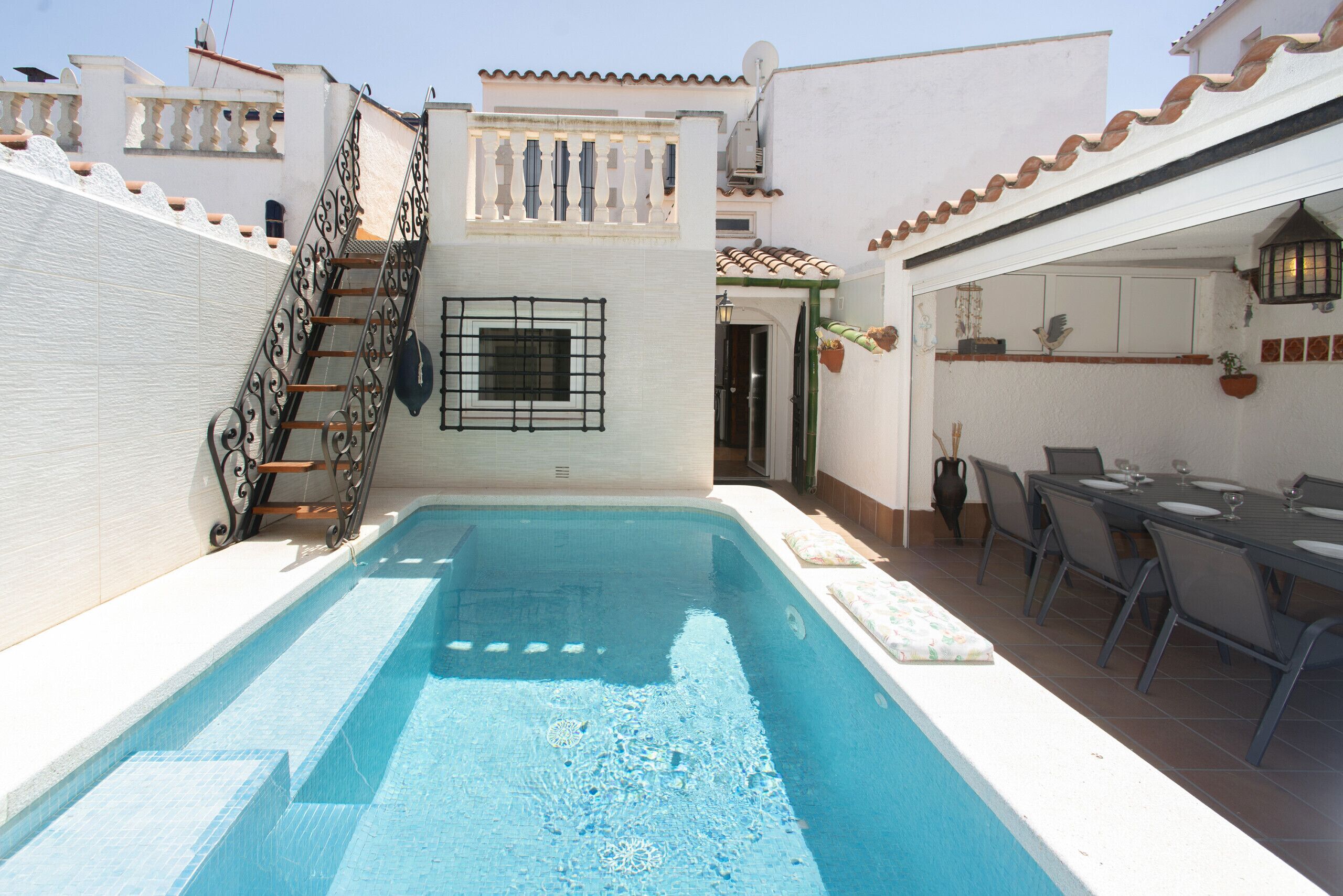 A swimming pool with a metal ladder, a dining area with a table and chairs, and a white building with a tiled roof.