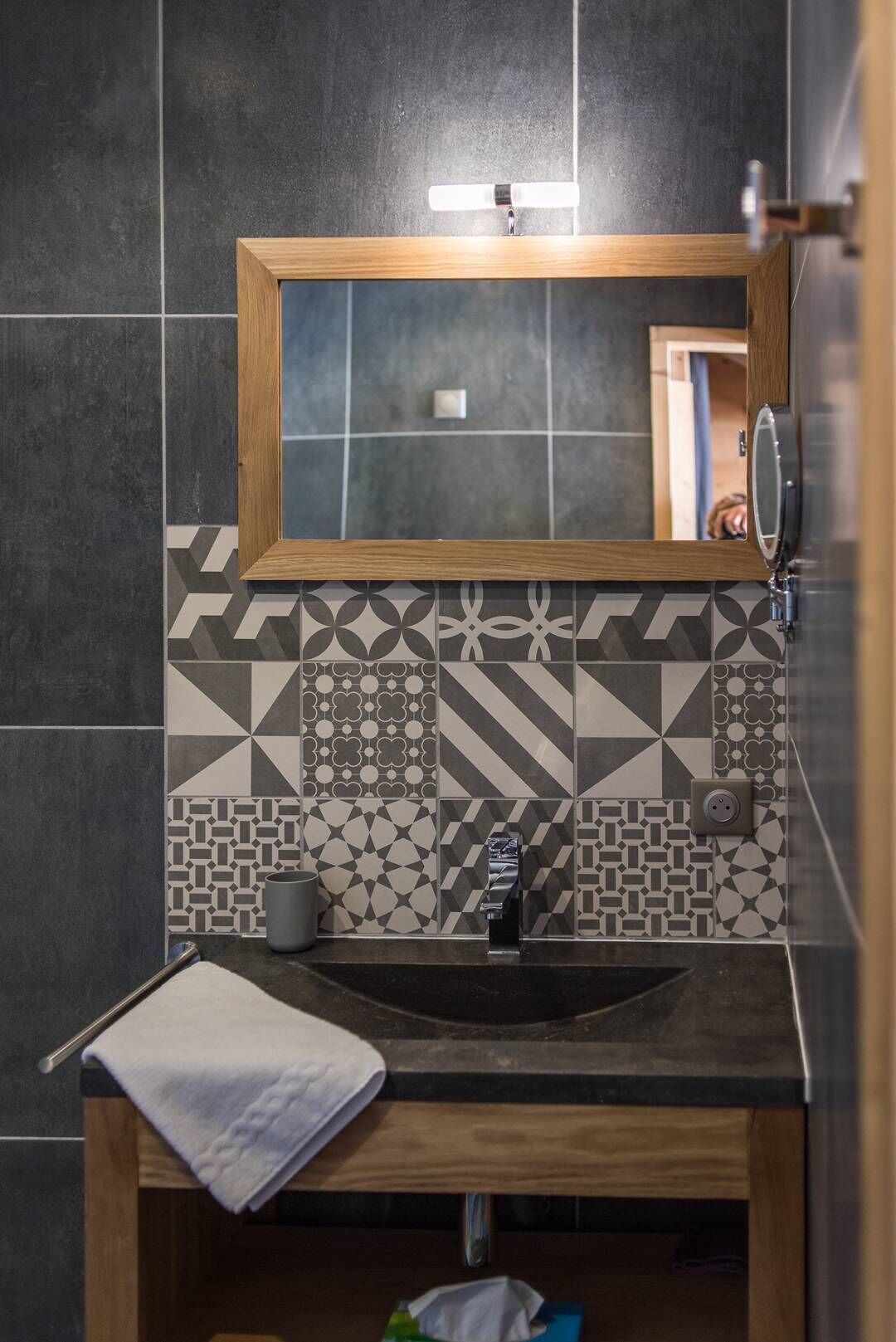 Une salle de bain avec un meuble-lavabo en bois, un miroir et un lavabo. Le mur est recouvert de carreaux aux motifs géométriques.