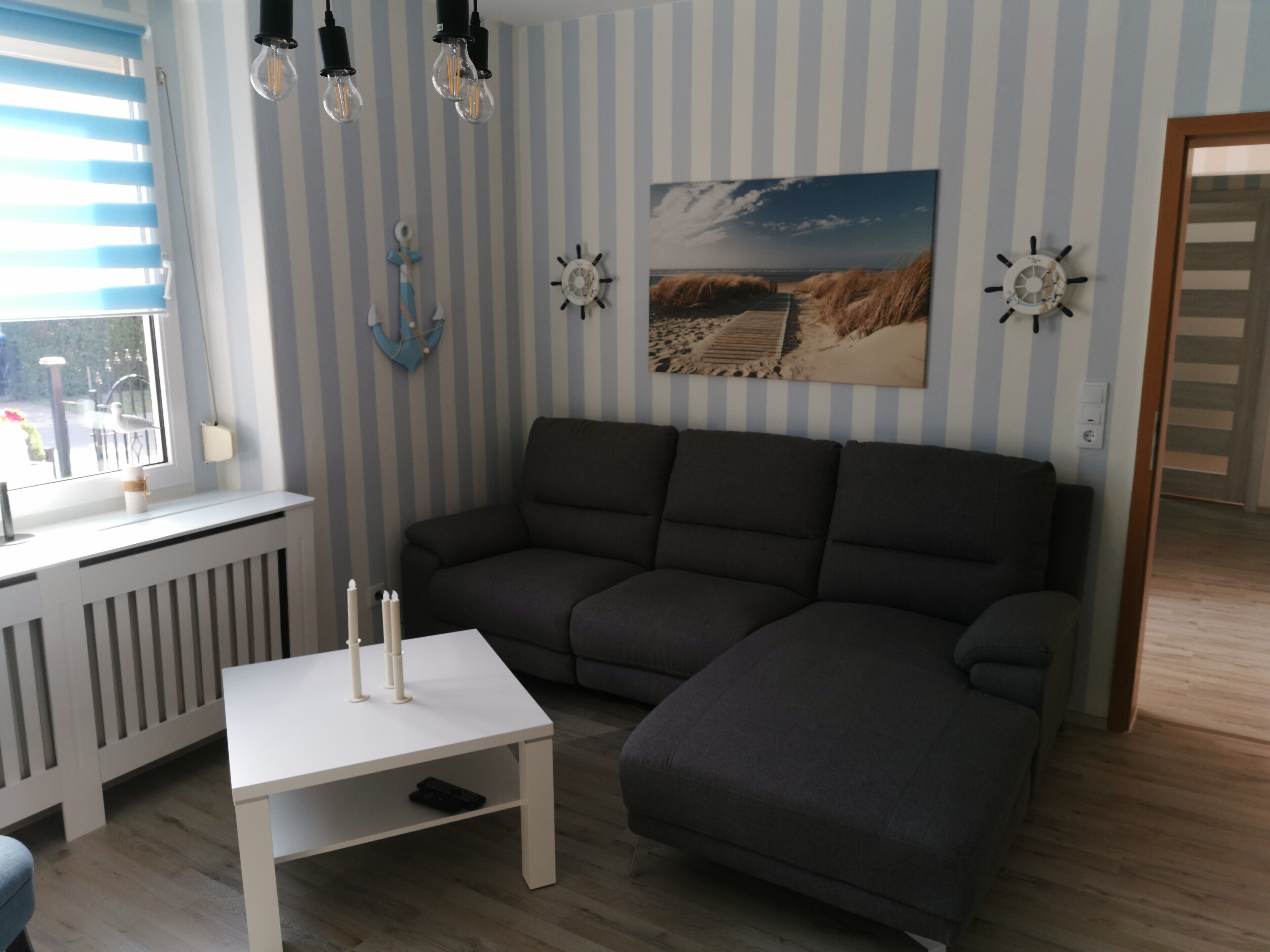 Uma sala de estar com sofá seccional, mesa de centro branca, janela com persianas e um relógio de parede.