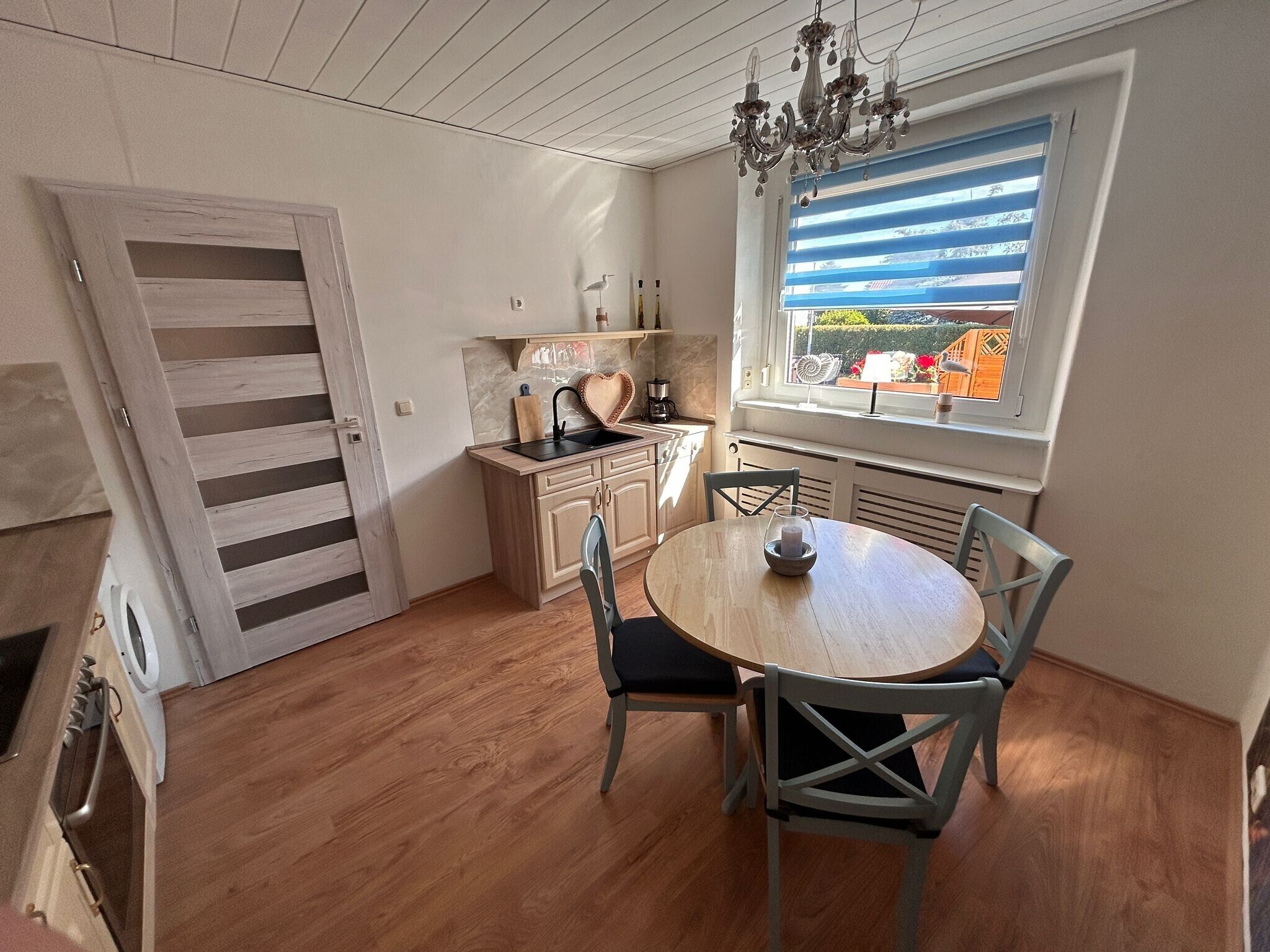 Een keuken met een houten vloer, een eettafel met stoelen, een raam met zonwering en een kroonluchter.