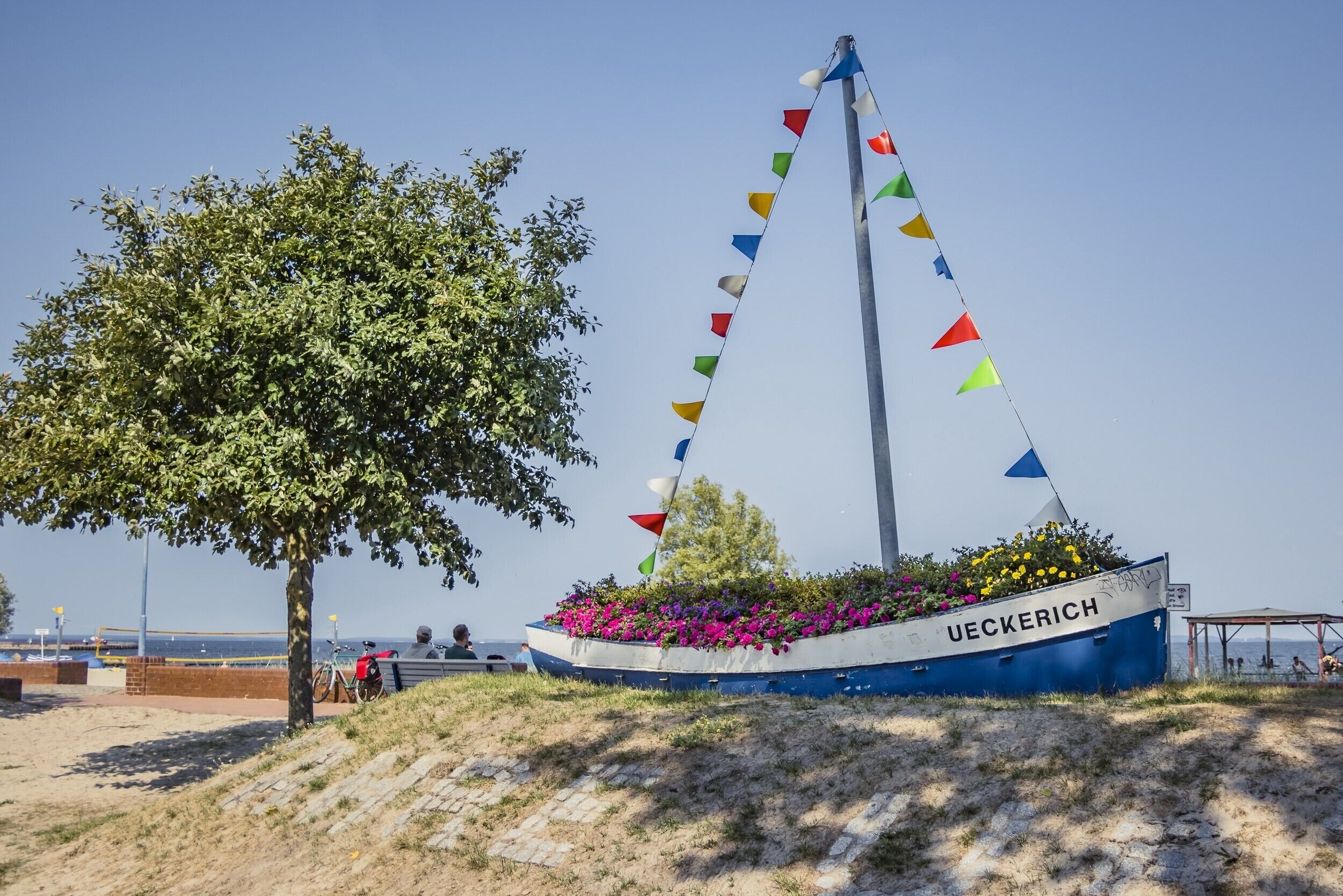 Ein Boot mit der Aufschrift 'UECKERICH', verziert mit bunten Wimpeln, umgeben von einer grünen Wiese mit einem Baum.