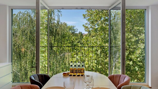 Comedor con una mesa dispuesta para cuatro, ventanas amplias con vistas a árboles y una botella de agua sobre la mesa.