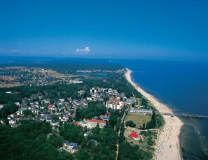 Vista aérea de uma cidade costeira com praia, áreas residenciais e um edifício de destaque com telhado vermelho.