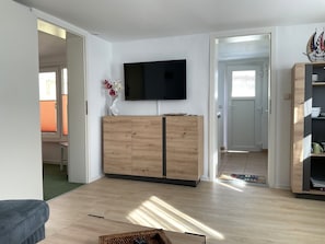 Un salon moderne avec un téléviseur à écran plat fixé au mur, un meuble en bois, un canapé en osier et une porte donnant sur une autre pièce.