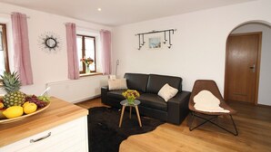 A living room with a black sofa, a wooden coffee table, a wicker chair, a black rug, and a fruit bowl on a wooden cabinet.