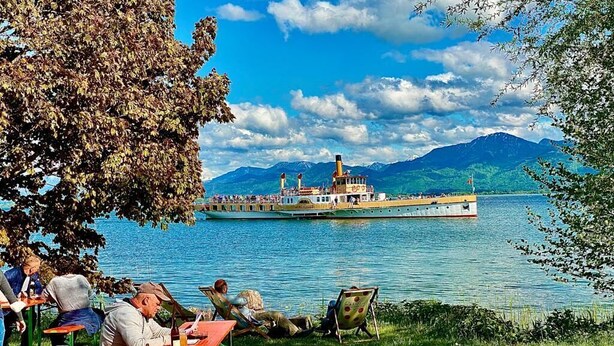 A lakeside setting with people relaxing at tables, a steamboat in the background, and mountains in the distance.