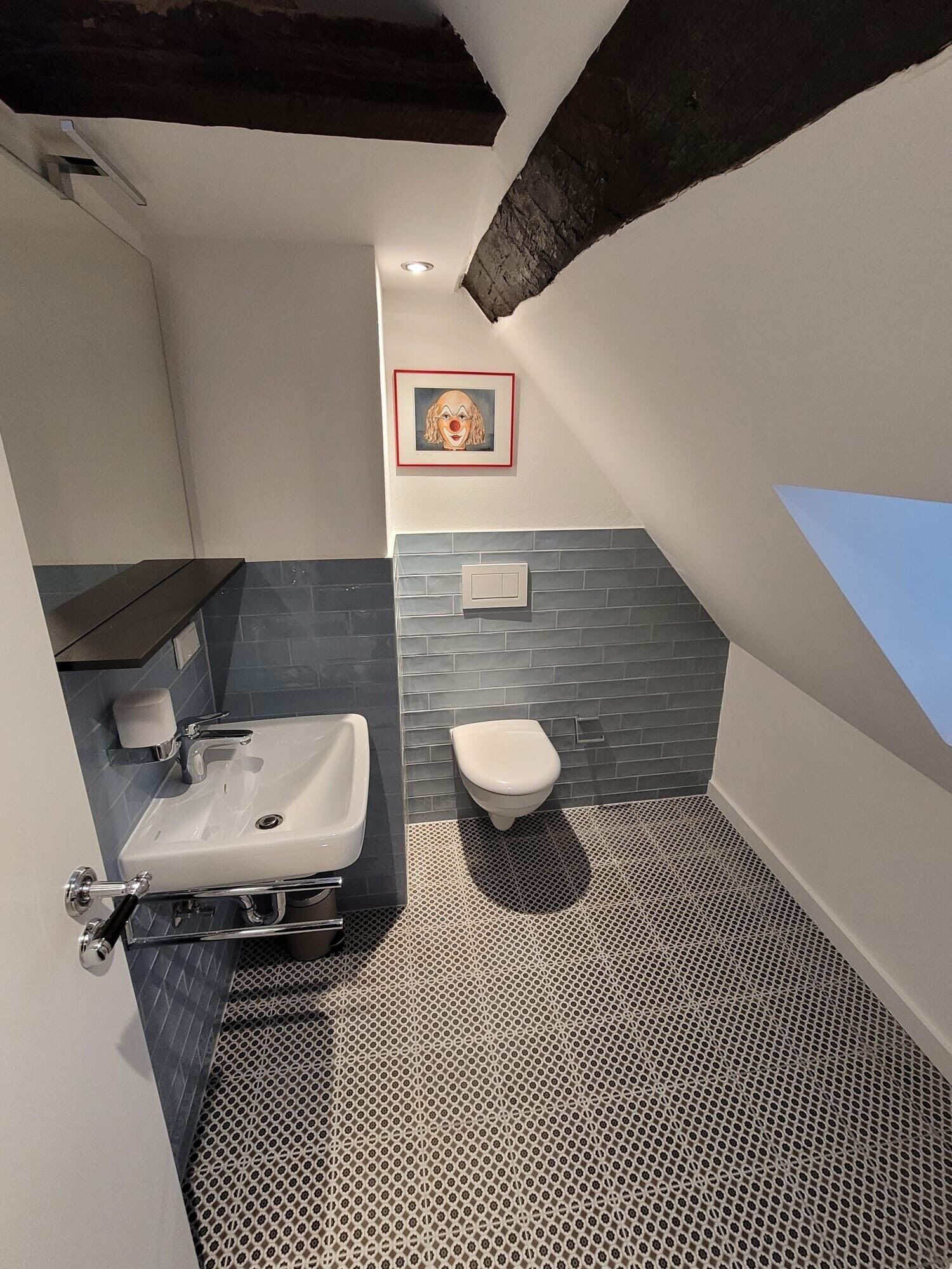 Un baño moderno con lavamanos blanco, inodoro y zona de ducha. Hay un cuadro enmarcado en la pared y una ventana pequeña.