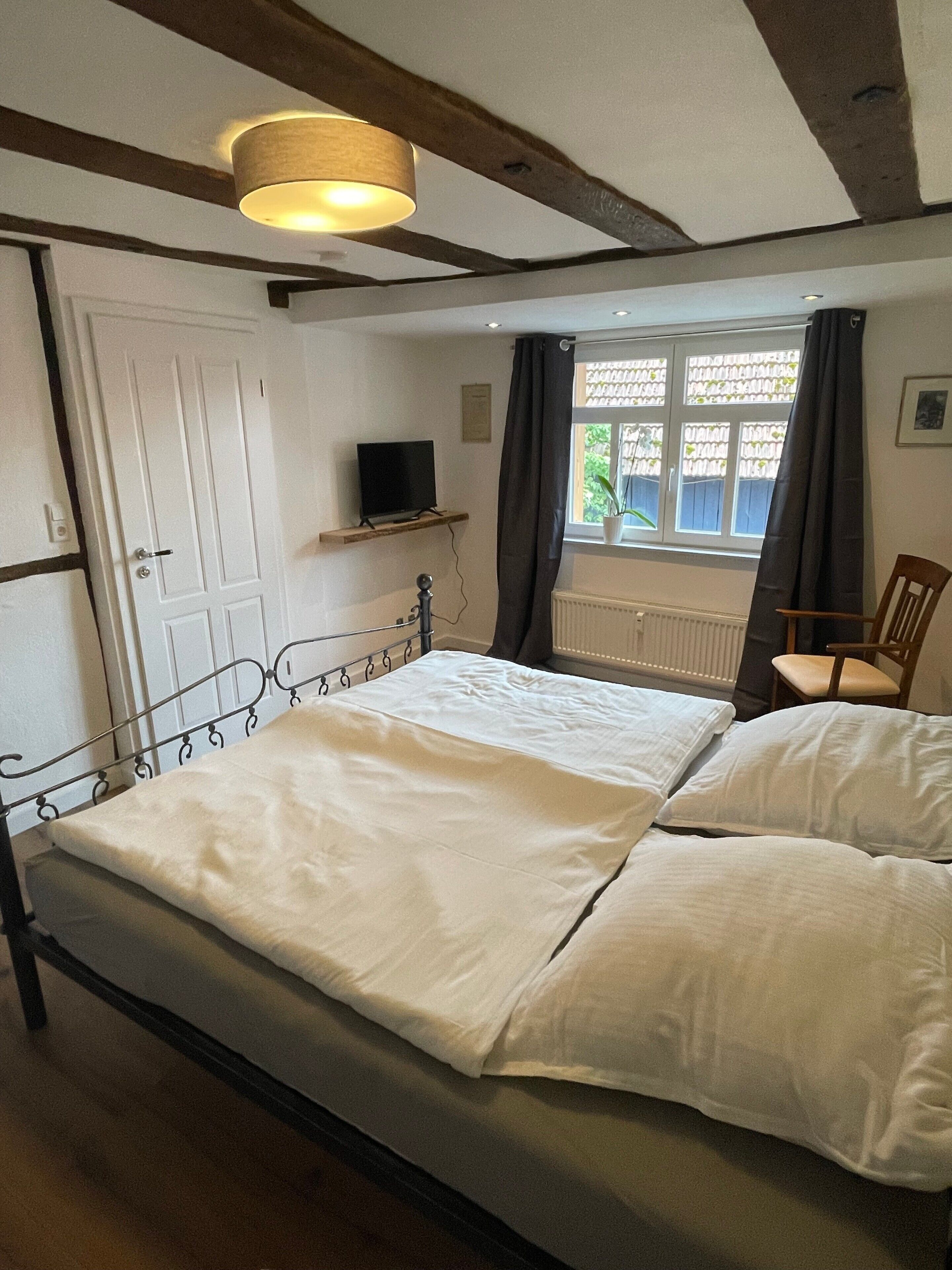 A bedroom with a bed, a television on a shelf, a window with curtains, and a chair.