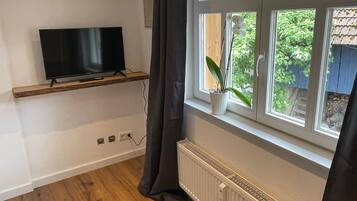Una habitación con una televisión sobre un estante, una ventana que ofrece vistas al verdor, y un radiador.