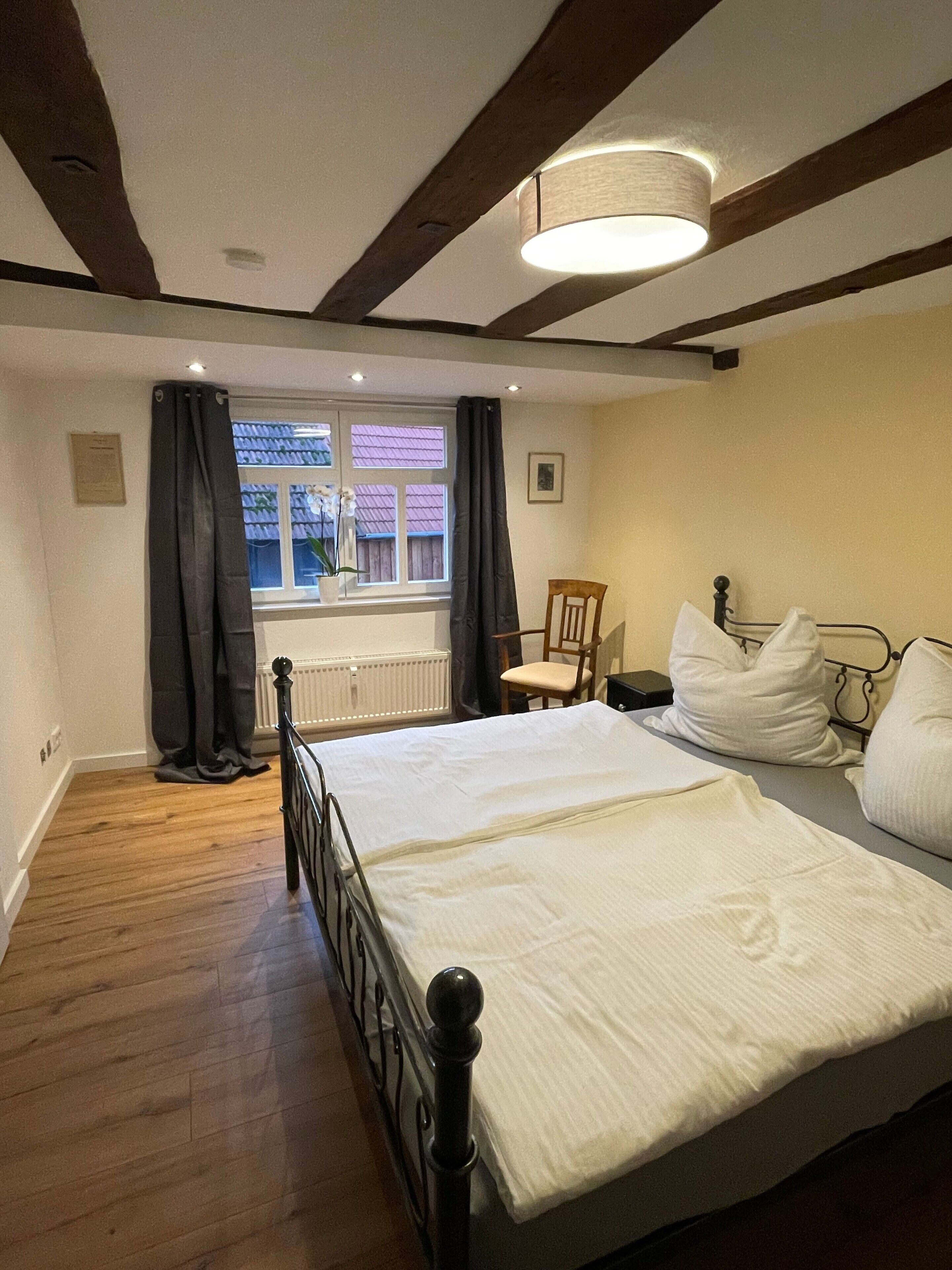 Une chambre à coucher avec un lit, une chaise, une fenêtre avec des rideaux et un luminaire au plafond.