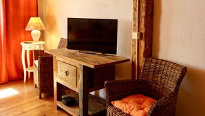 Une pièce avec un plancher en bois, un téléviseur posé sur un meuble en bois, une chaise en osier avec un coussin orange, une petite table blanche avec une lampe, et un rideau rouge.