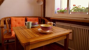 Une table en bois, deux tasses et soucoupes à motifs, une chaise avec un coussin orange et une fenêtre avec des plantes en pot.