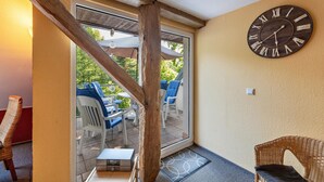 Une pièce avec un plafond en bois, une grande horloge murale, une porte-fenêtre coulissante donnant sur un espace extérieur aménagé avec des chaises et des tables, et un fauteuil en osier à l’intérieur.