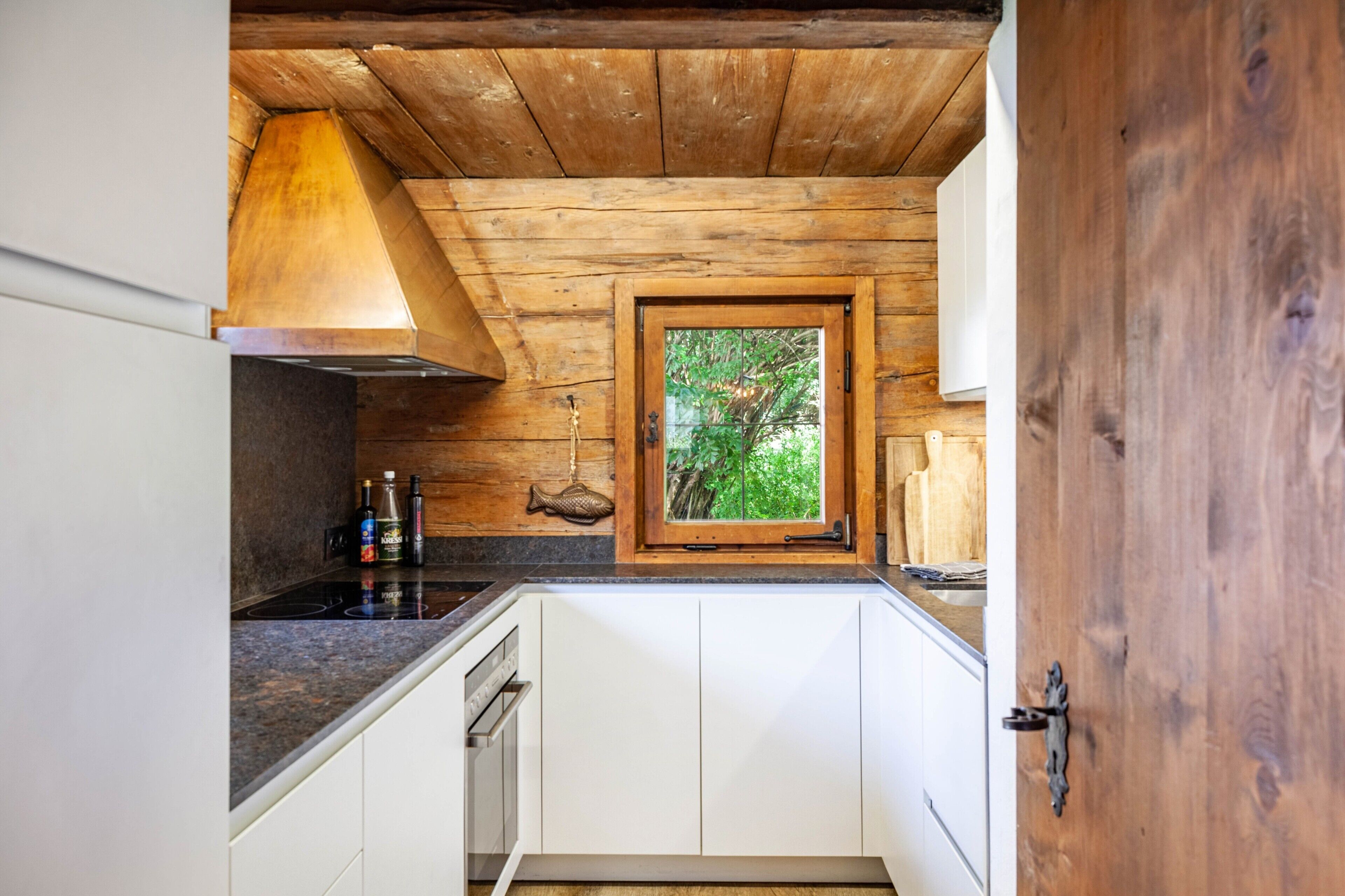 Eine Küche mit Holzbalkendecke, einem Fenster mit Blick ins Grüne, weißen Schränken und einem Edelstahlherd.