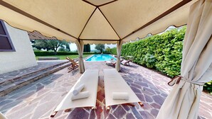 Un espacio al aire libre techado con suelo de piedra, dos sillones de descanso y una piscina al fondo.