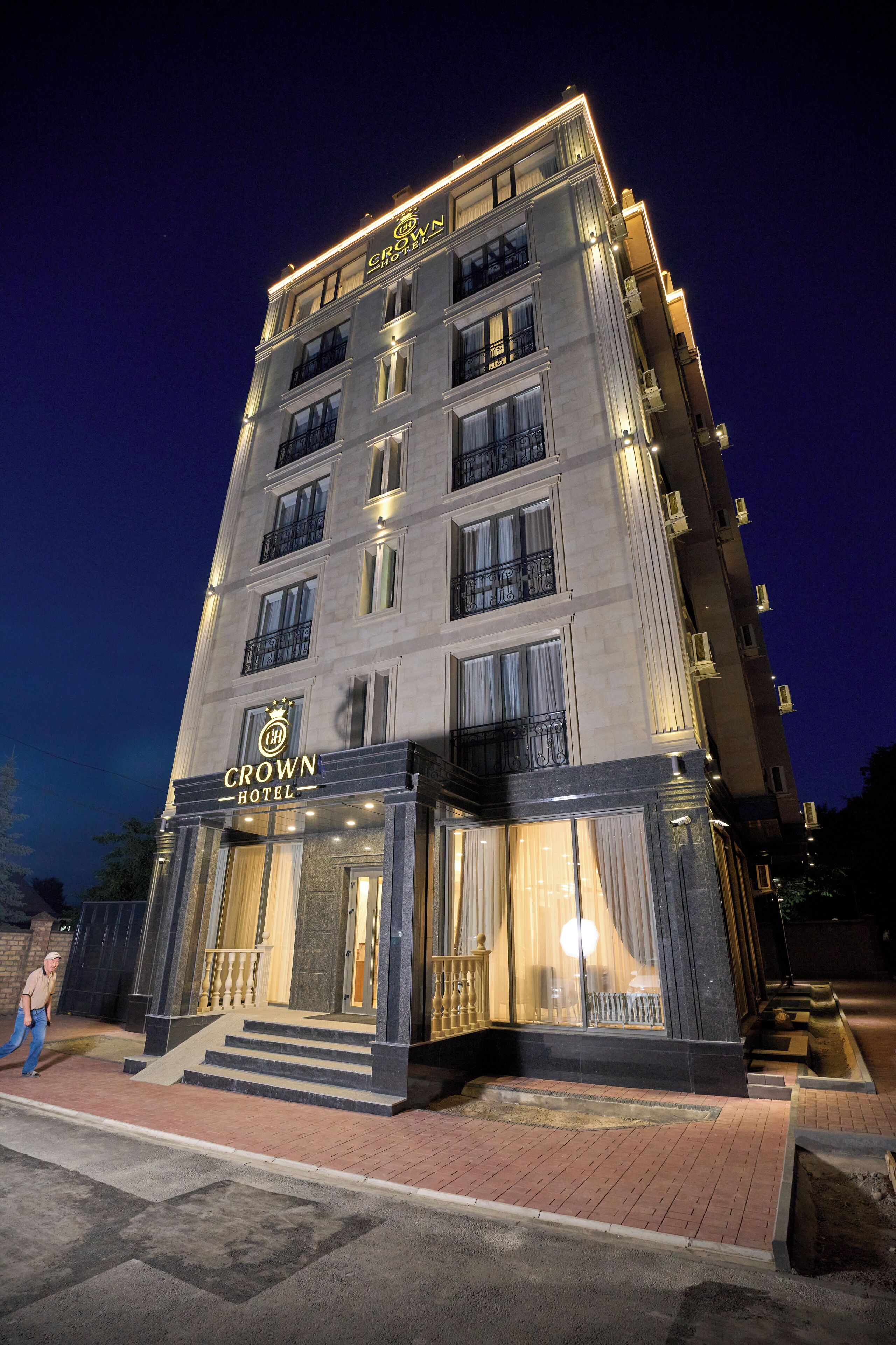 Un hôtel à plusieurs étages, reconnaissable à son grand panneau « Crown Hotel », doté d’une entrée imposante avec des marches et un balcon.