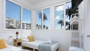 Een hotelkamer met twee bedden, een raam met uitzicht op palmbomen en gebouwen, een witte kast en een ladder.